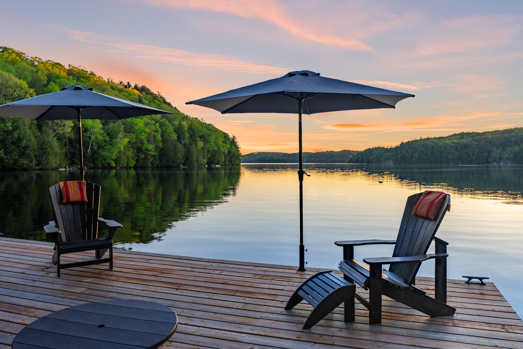 Two Adirondack chairs and umbrellas on the dock framing a glassy sunset lake