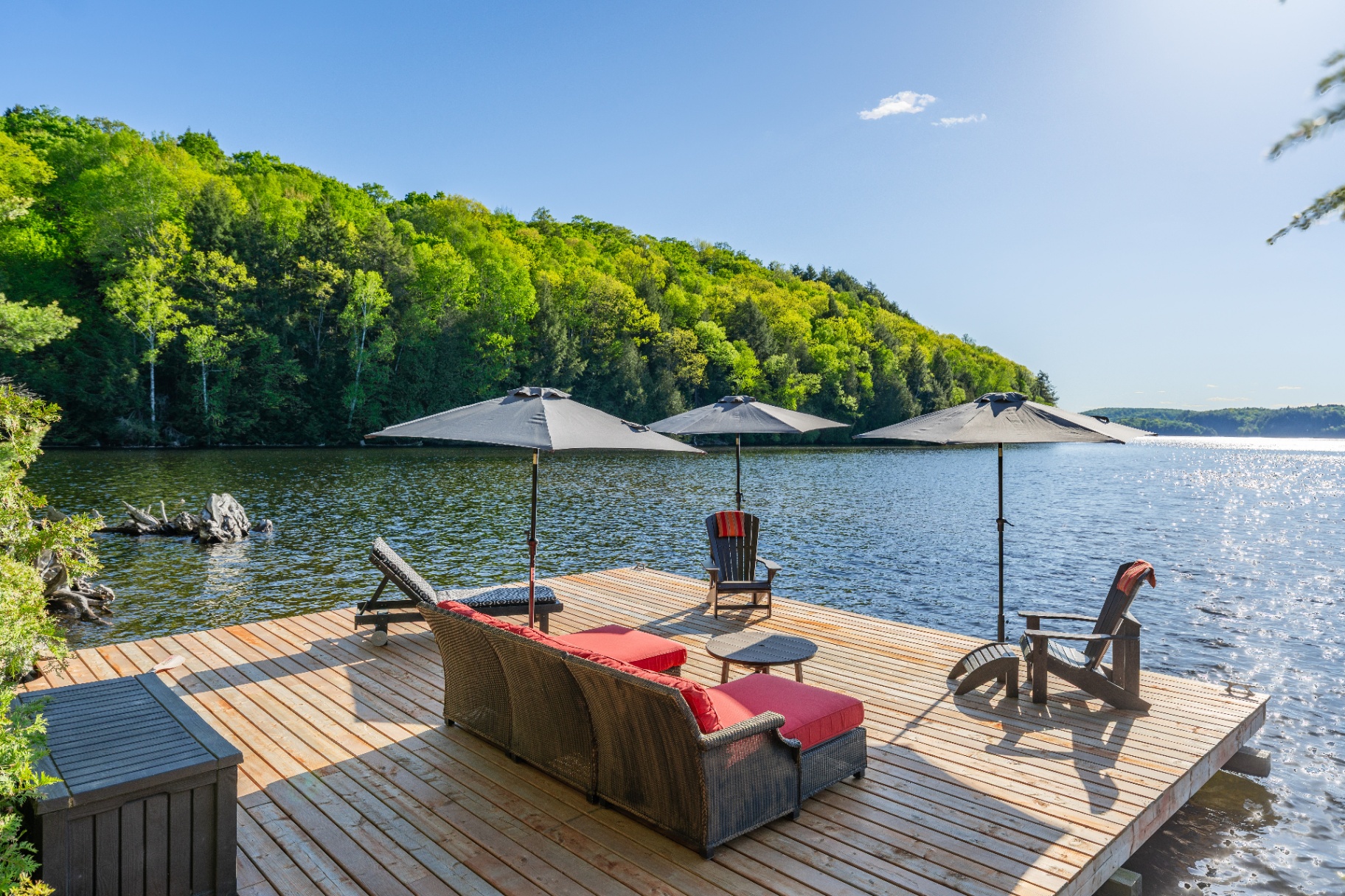 Sun-dappled dock with red lounger, Adirondack chairs, and umbrellas over open lake
