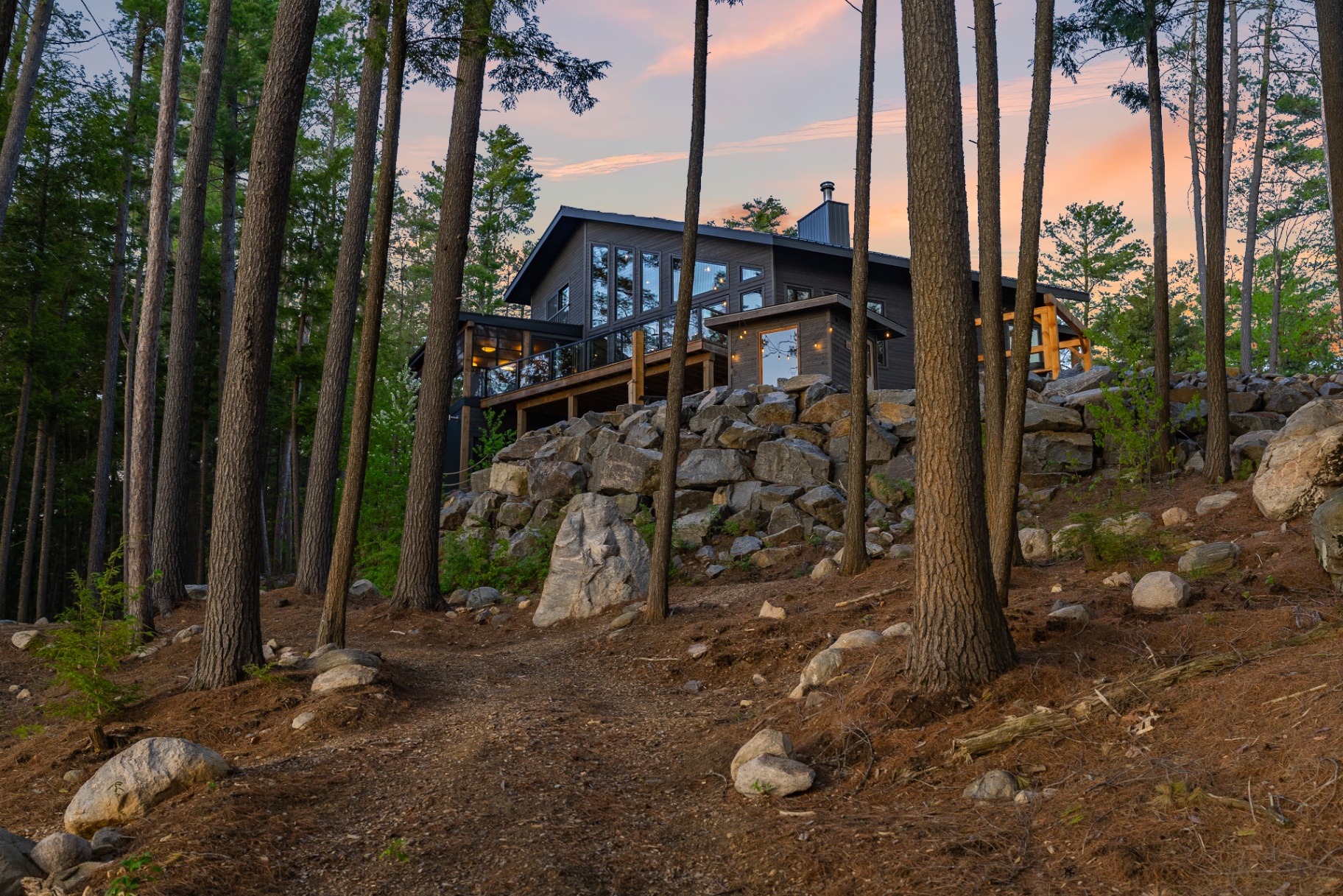Rear of the house seen through tall pines with sauna, deck, and warm sunset sky